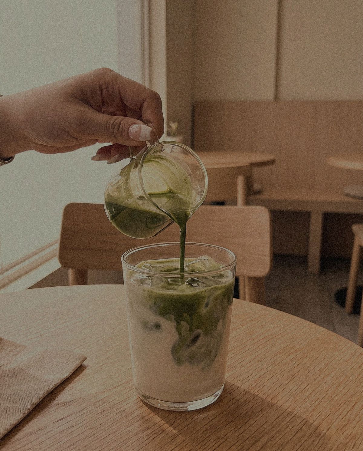 A hand pouring green matcha from a small glass jug into a tall glass of iced milk on a wooden cafe table