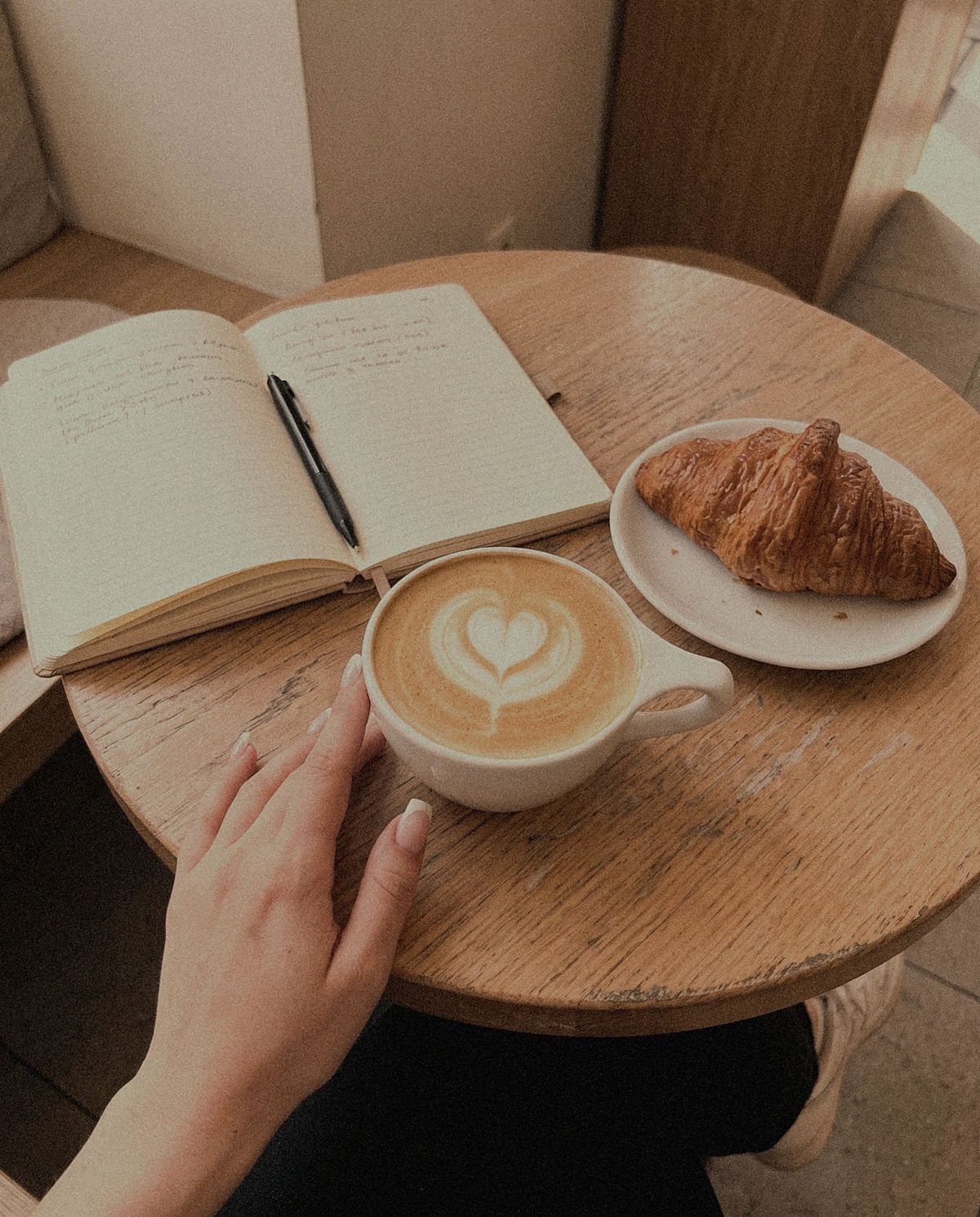 Overhead view of a round wooden cafe table with an open handwritten notebook and pen, a latte with heart latte art, and a croissant on a small plate, with one hand resting on the cup