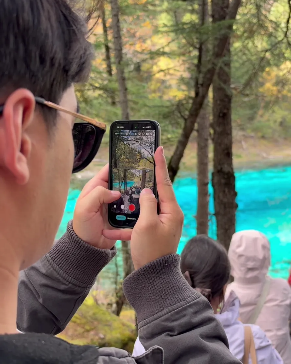 Over-the-shoulder view of a man holding up his phone to film a bright turquoise pool through pine trees, the ContentCam split preview on screen