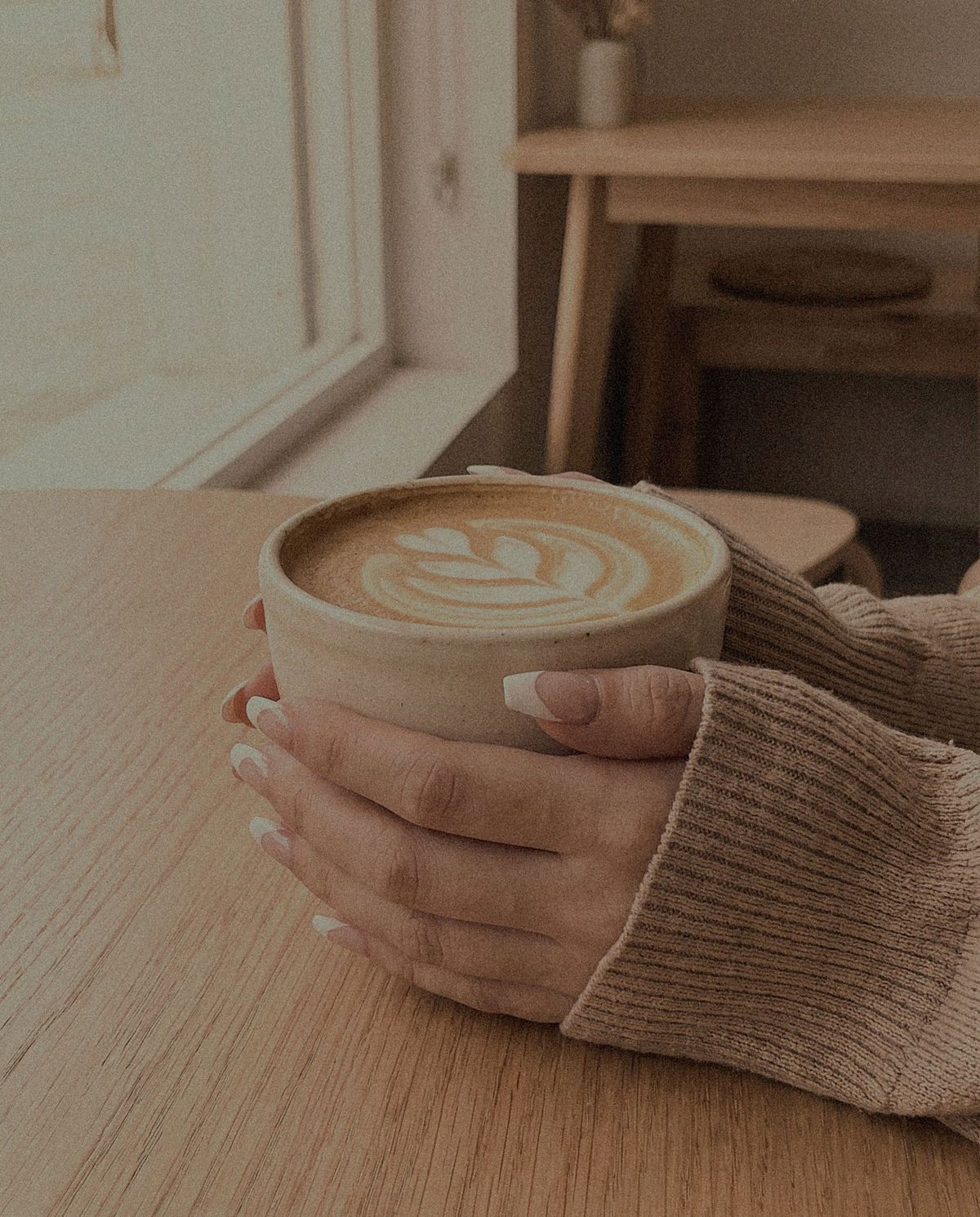 Close detail shot of two hands in knit sleeves wrapped around a ceramic cup of latte on a wooden table
