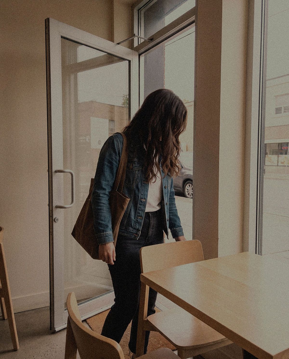 A low, wide shot from table height of a woman in a denim jacket walking in through a glass cafe door, her face turned away from the camera