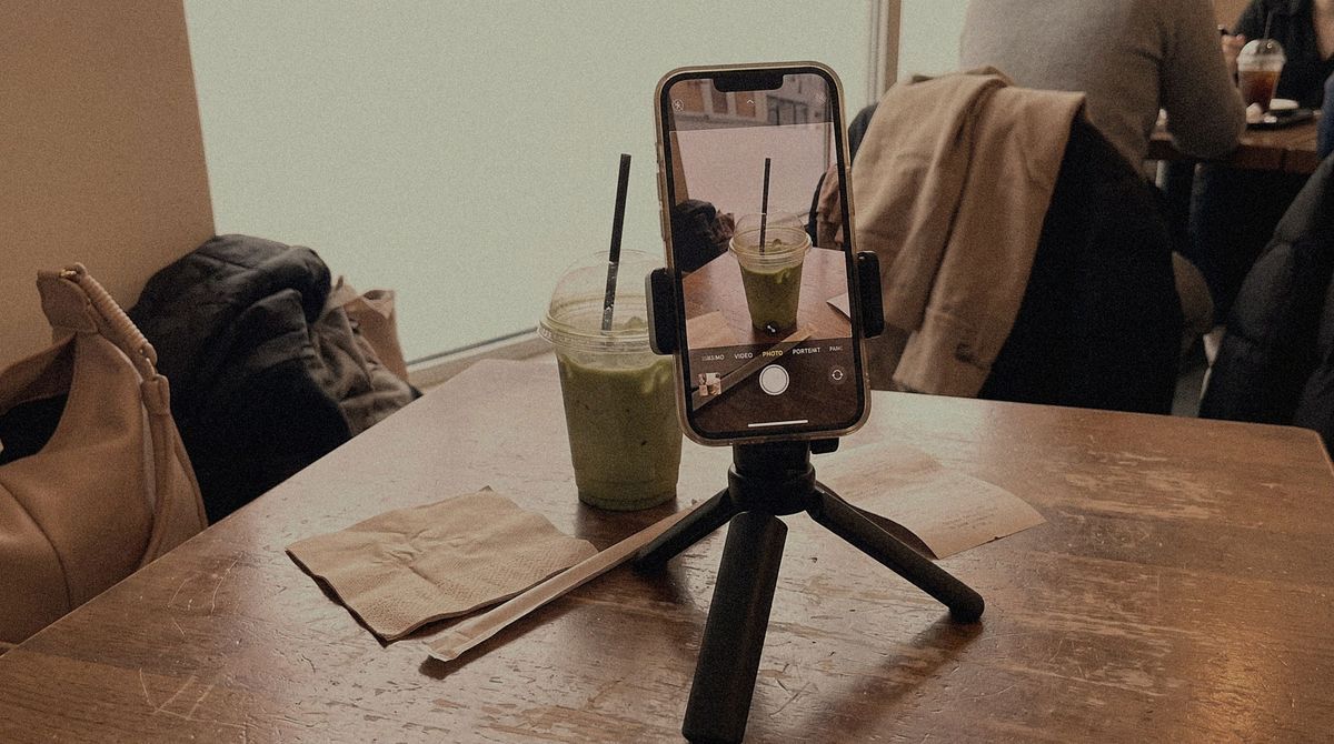 A phone in a clamp on a small black tabletop tripod at a cafe table, its screen showing the camera framing an iced matcha beside it